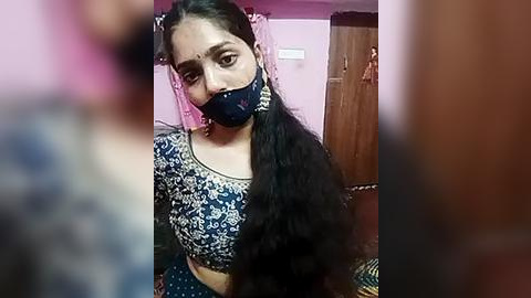 Media: A video of a woman with medium-dark skin and long black hair in a traditional blue and white patterned blouse, wearing a black face mask, standing indoors with pink walls and wooden furniture.