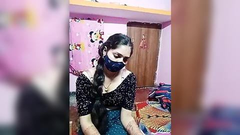 Media: Video of a woman with dark hair in a braid, wearing a black and white polka dot dress and blue face mask, sitting on a colorful rug in a pink room with cartoon curtains and a wooden door.