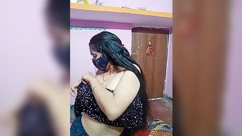 Media: A video of a plus-sized woman with long black hair, wearing a black face mask and a black polka-dot top, adjusting her bra in a dimly lit, cluttered bedroom with a wooden door and colorful bedding.