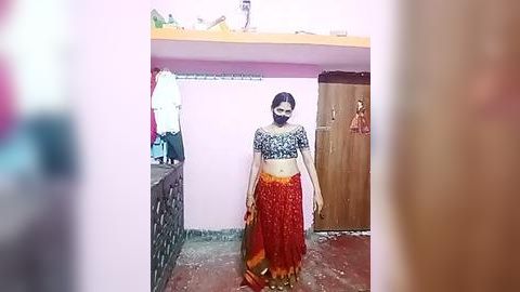 Media: Video of a young woman with medium skin tone, wearing a black crop top and vibrant orange saree, standing in a modest, cluttered room with pink walls and a wooden door.