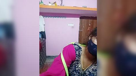 Media: Video of a woman lying on a bed in a small room with pink walls and a wooden cupboard, wearing a black face mask and a blue floral-patterned dress, with a yellow safety harness around her waist.