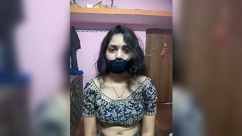 Media: Video of a South Asian woman with dark hair, wearing a blue face mask and a patterned crop top, in a dimly lit, cluttered room with pink walls and a wooden cabinet.