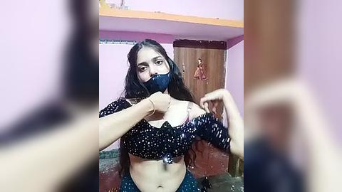 Media: Video of a young South Asian woman in a bedroom, wearing a dark blue mask, black crop top, and blue pants, adjusting her top, with blurred background.