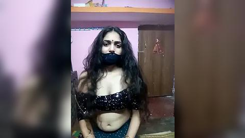 Media: Video of a South Asian woman with long black hair, wearing a black off-shoulder crop top, and a blue mask, in a modest, cluttered room with pink walls, wooden door, and hanging clothes.