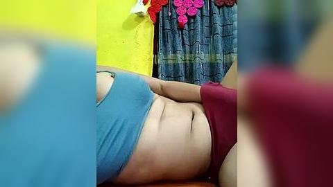 Media: Video of a woman with fair skin lying on a bed, wearing blue and red underwear, in a room with yellow and blue walls, floral curtains, and a yellow towel hanging.