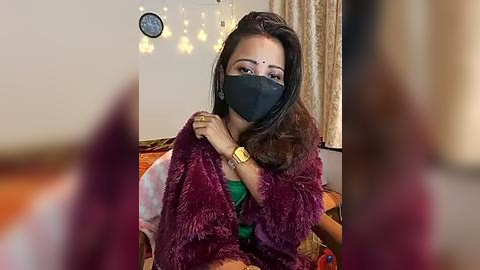 Media: Video of a woman with long dark hair, wearing a black face mask, burgundy shawl, and gold watch, seated on a couch in a cozy, dimly lit room with a clock and string lights in the background.