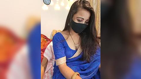 Media: Video of a young woman with long black hair, wearing a blue saree, black mask, and gold jewelry, in a dimly lit room with blurred figures.