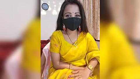 Media: Video of an Indian woman with medium skin tone, long dark hair, wearing a yellow saree, black mask, and gold jewelry, sitting indoors, blurred background.