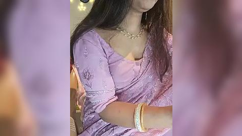 Media: A video of a woman in a lavender saree with intricate embroidery, wearing gold jewelry and a necklace, sitting indoors.