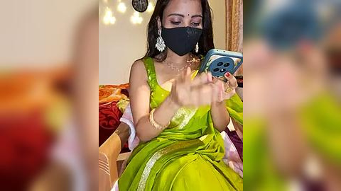 Media: Video of a South Asian woman in a green sari with a black face mask, gold earrings, and a phone in hand, sitting on a red sofa with a blurred background.