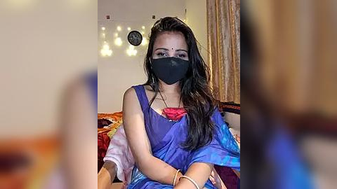 Media: Video of a young woman with long black hair, wearing a blue saree, black mask, and red blouse, sitting on a bed with a colorful blanket. Background includes a wall clock and curtains.