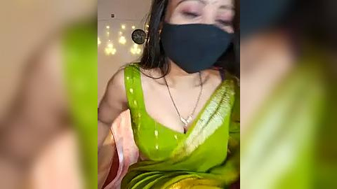 Media: A video of a South Asian woman with medium skin tone, wearing a bright green sari, black face mask, and gold earrings. She has a slim physique and is indoors with warm lighting.