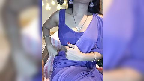 Media: Video of a woman in a blue saree, with long earrings, adjusting her blouse, wearing a necklace, and bracelets, sitting on a bed with a blurred background.