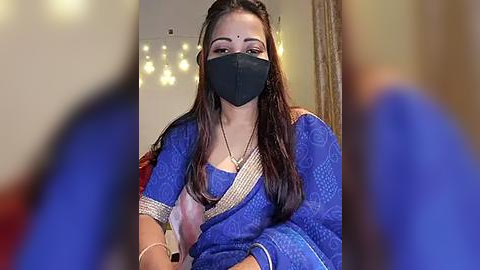 Media: Video of a South Asian woman with long brown hair, wearing a blue saree with intricate patterns, a black face mask, and a gold necklace. The blurred background features fairy lights and a beige curtain.