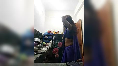 Media: Video of a woman in a purple saree and blue blouse, adjusting her hair in a small, cluttered bathroom with a sink, mirror, and a shelf filled with personal items.