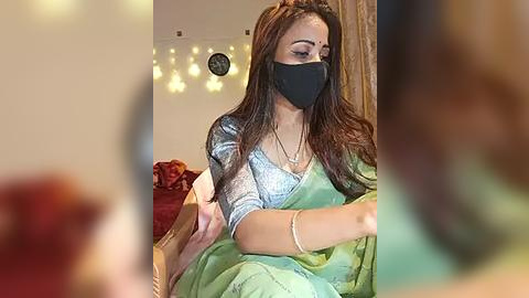 Media: Video of a South Asian woman in a green saree, black mask, and light blue blouse, seated indoors with blurred background.