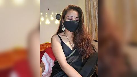 Media: Video of a young woman with long brown hair, wearing a black mask, black dress, and silver earrings, seated indoors with a blurred background.