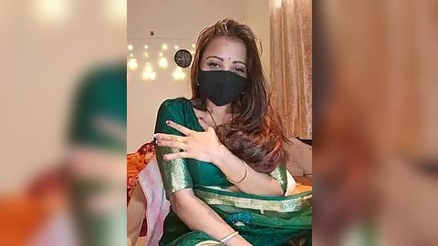 Media: Video of a woman in a green sari with a gold border, wearing a black face mask, sitting indoors, blurred background.