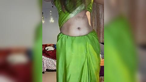 Media: Video of a light-skinned person wearing a green, see-through saree, revealing their bare midriff and partially covered breasts. The background shows a cluttered living room with a bed, stuffed toys, and a hanging lamp.