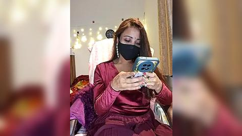 Media: Video of a woman with long brown hair, wearing a black face mask, maroon top, and matching pajamas, sitting in a chair, looking at her smartphone. Background includes a blurred, warmly lit room with fairy lights.