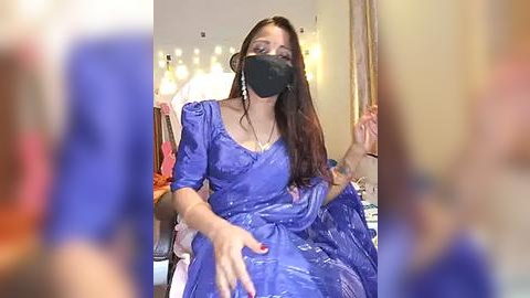 Media: Video of a woman in a blue saree with a mask, sitting in a room with soft, warm lighting and blurred people in the background.