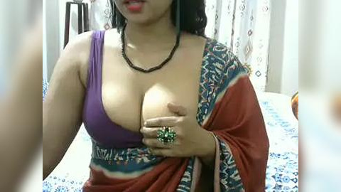 Media: A video of a South Asian woman with medium skin tone, wearing a purple bra and an orange sari with intricate blue patterns. She holds her breast with a green ring, in a bedroom setting.