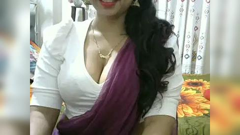 Media: Video of a South Asian woman with long, wavy black hair, wearing a white blouse and a purple sari, smiling, in a brightly lit room with patterned curtains and a floral cushion.