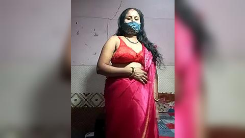 Media: Video of a woman in a red saree with a blue face mask, standing in a dimly lit room with peeling wallpaper and a patterned tile floor.