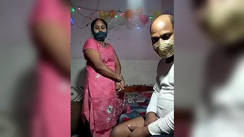 Media: A video shows a woman in a pink sari, mask, and man in a white shirt, praying in a dimly lit room with string lights and patterned wall.