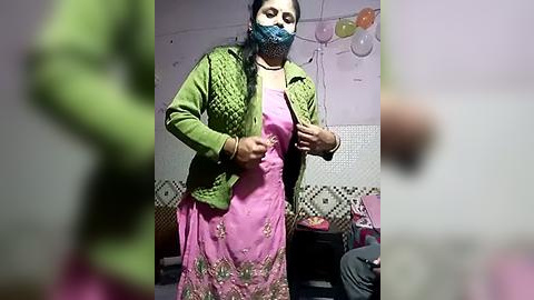 Media: A video of an Indian woman in a green jacket over a pink sari with gold embroidery, wearing a blue mask, adjusting her mask in a room with white walls and a patterned carpet.