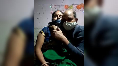 Media: Video of two masked healthcare workers in green scrubs comforting an elderly woman wearing a black mask and green sari. Background features festive orange and green lights.