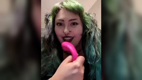 Media: A close-up video of a woman with green hair and dark makeup licking a pink dildo. She has a medium skin tone and is indoors, with blurred surroundings.