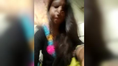 Media: Video of a dark-skinned woman with long, curly hair wearing a colorful, patterned shawl over a black shirt, captured in a blurry, out-of-focus setting.