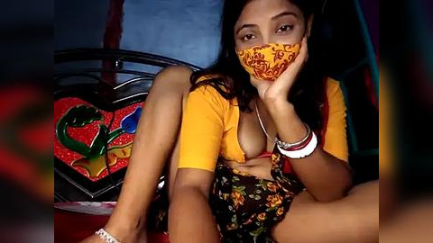 Media: Video of a South Asian woman with medium skin tone, wearing a yellow blouse and floral skirt, sitting on a colorful, patterned cushion. She has long black hair and is masked, resting her chin on her hand.