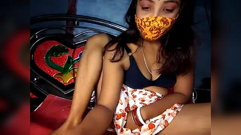 Media: Video of a young woman with medium brown skin, wearing a yellow face mask, black bra, and orange-patterned skirt, sitting on a bed with red and green heart decorations, in a dimly lit room.