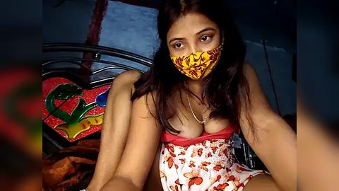 Media: Video of a young South Asian woman with medium-brown skin, long dark hair, and wearing a red and yellow floral-patterned face mask, sitting on a bed with colorful, cartoon-patterned pillows.