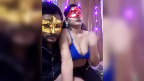 Media: Video of a woman with a blue bra and red mask, sitting next to a man with a gold mask, both in a dimly lit room with purple string lights.