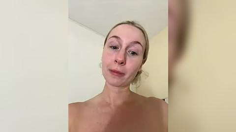 Media: Video of a topless, fair-skinned, blonde woman with a slight smile, in a simple indoor setting with cream-colored walls and a ceiling.