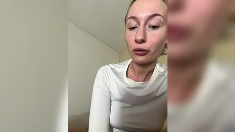 Media: Video of a fair-skinned, blonde-haired woman with arched eyebrows, wearing a white long-sleeved top, looking slightly to the side, with blurred background.