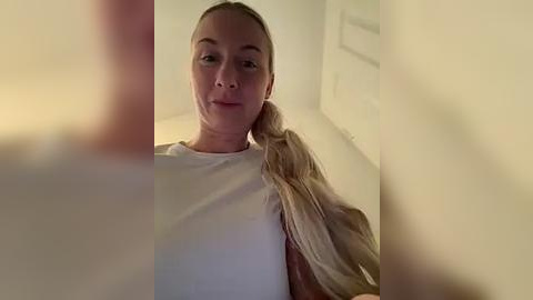 Media: Video of a blonde woman with a ponytail, wearing a white t-shirt, sitting in a dimly lit room with beige walls and a door in the background.