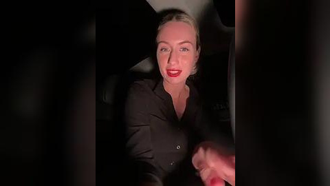 Media: Video of a blonde woman with fair skin, wearing a black shirt, seated in a car at night. The image is dimly lit, with shadows obscuring parts of her face and surroundings.