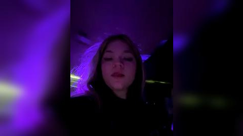 Media: Video of a young woman with fair skin and long brown hair, wearing a black outfit, illuminated by purple and blue lights, creating a moody, nightclub atmosphere.