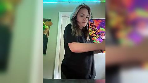 Media: A blurry video of a young woman with wavy, light brown hair, wearing a black t-shirt, standing in front of a mirror in a colorful room with a vibrant, psychedelic mural on the wall.