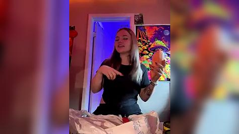 Media: Video of a young woman with long blonde hair, wearing a black t-shirt, holding a phone, in a brightly lit room with colorful wall art and a red chair.