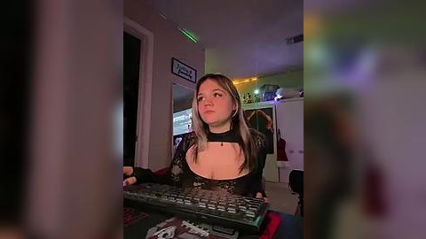 Media: Video of a young woman with light skin and straight, shoulder-length brown hair, wearing a black, lace-trimmed top, focused on a computer keyboard. Background shows a dimly lit room with neon lights, cluttered with gaming equipment.