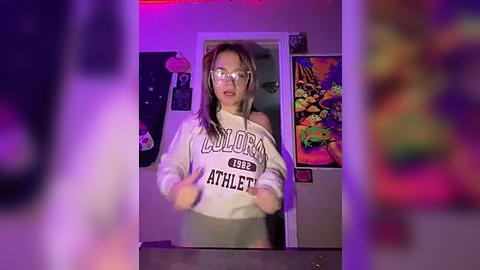 Media: Video of a young woman with glasses, wearing a \"Loudon Athletics\" sweatshirt and glasses, in a room with psychedelic posters, purple lighting, and a blurred figure in the background.