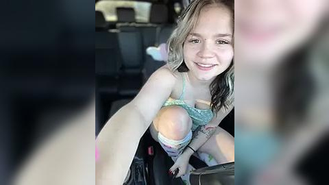 Media: Video of a young woman with light skin and blonde hair, wearing a colorful, low-cut top, sitting in a car. She smiles, and the car's interior is visible in the background.