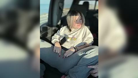 Media: Video of a young woman with shoulder-length blonde hair, wearing a white hoodie and gray pants, seated in a car with blurred background.