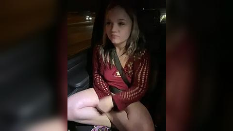 Media: Video of a young girl with light skin, long brown hair, and a red cardigan, sitting in a dimly lit car with blurred figures in the background.