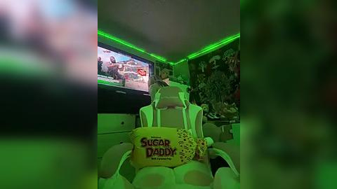 Media: Video of a cozy, dimly lit room with green LED lighting, featuring a white gaming chair, a yellow \"Sugar Daddy\" pillow, and a TV showing a cartoon, with a fish tank in the background.